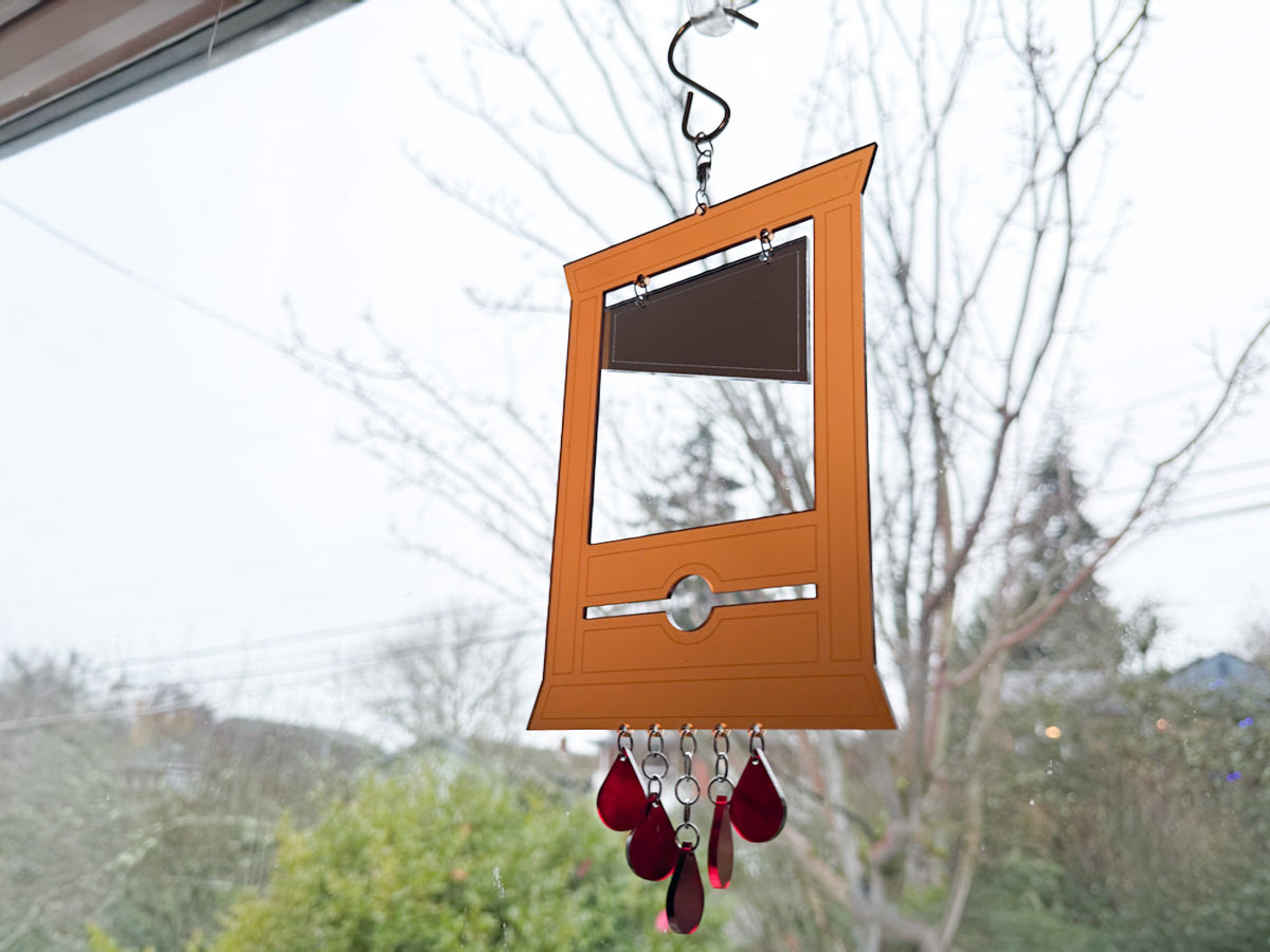 Handmade golden amber acrylic guillotine suncatcher hanging from S-hook in front of a window with bare winter trees, red acrylic teardrop blood drop dangles at bottom