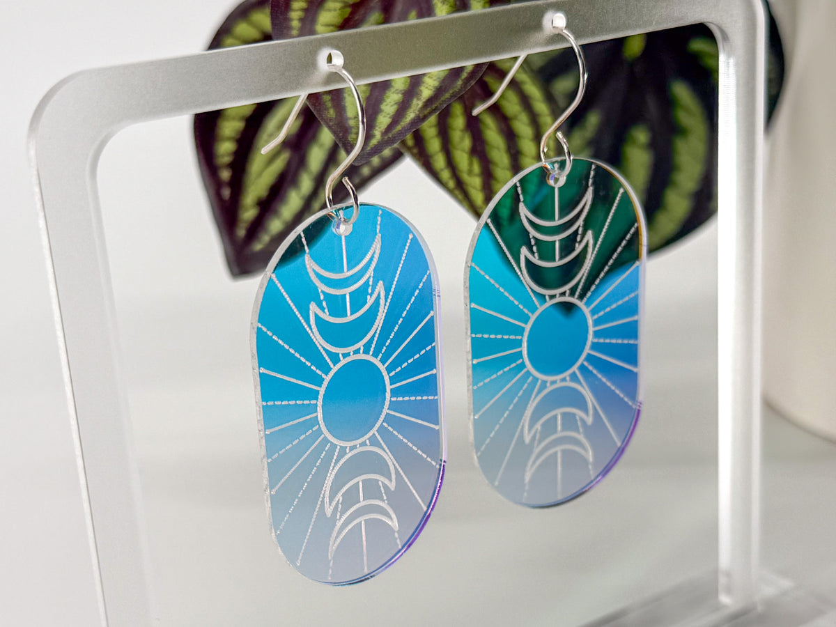 A pair of oval drop earrings with a engraved crescent moon and sun design, featuring a blue-to-purple gradient background, hanging on a clear acrylic display stand.