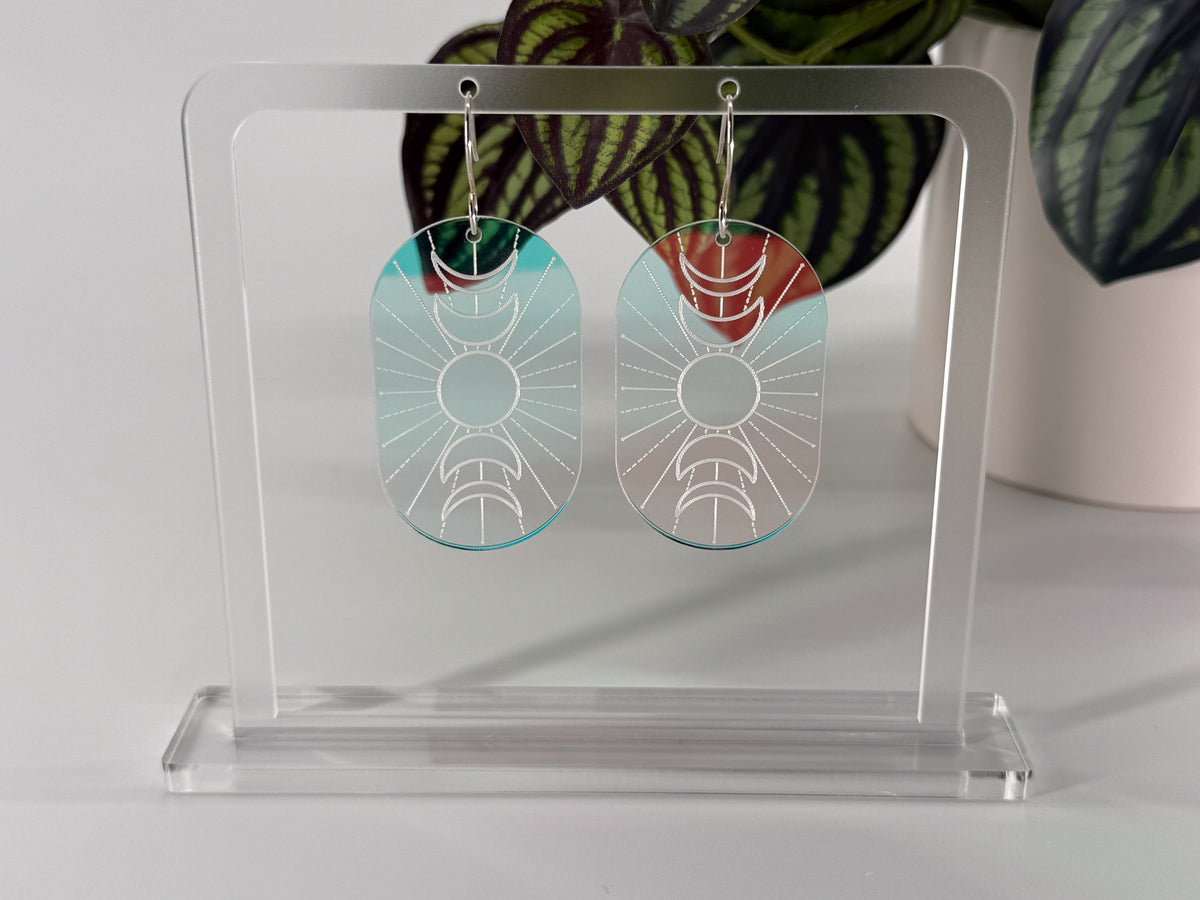 A pair of oval drop earrings with a sunburst and moon geometric design, displayed on a clear acrylic stand. The earrings feature a light blue-green gradient on one side and a white-to-clear gradient on the other, suspended from silver hooks.