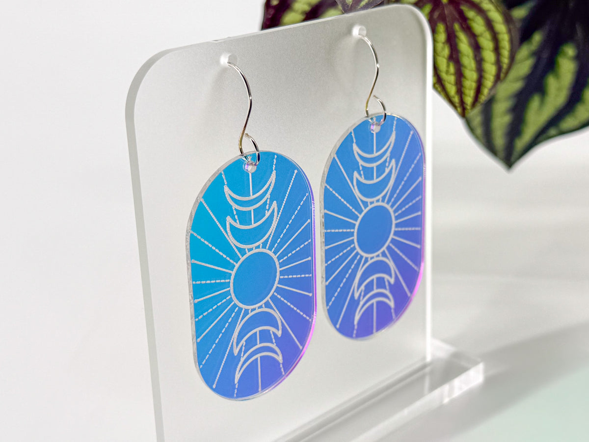 A pair of oval dangling earrings displayed on a clear acrylic stand. The earrings show a blue-to-purple iridescent  gradient background with an engraved moon phase design and silver hooks.