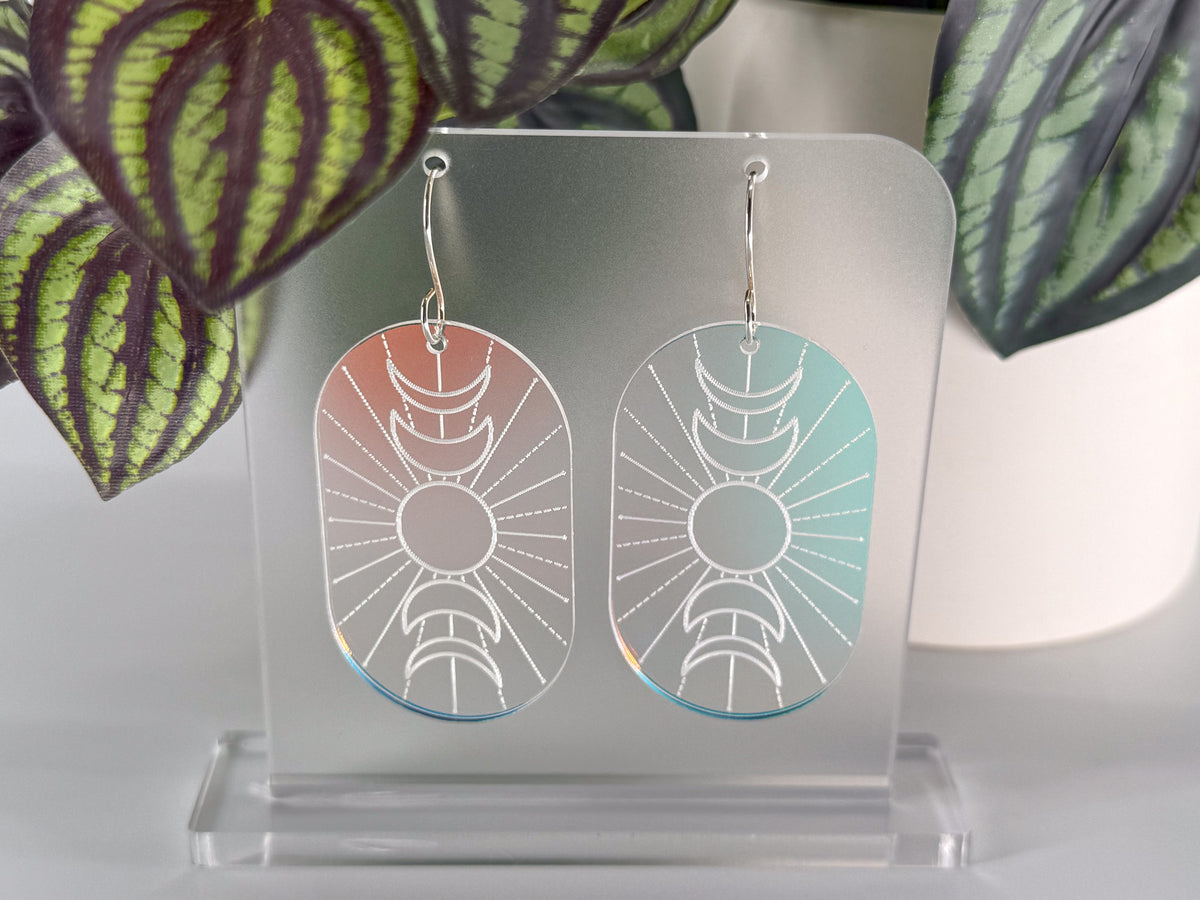 A pair of oval dangling earrings displayed on a clear acrylic stand, with a silver-toned metal hook and a white whimsigothic sunburst design. The left earring has a soft pink-to-blue iridescent gradient, and the right earring has a soft blue-to-green iridescent gradient.