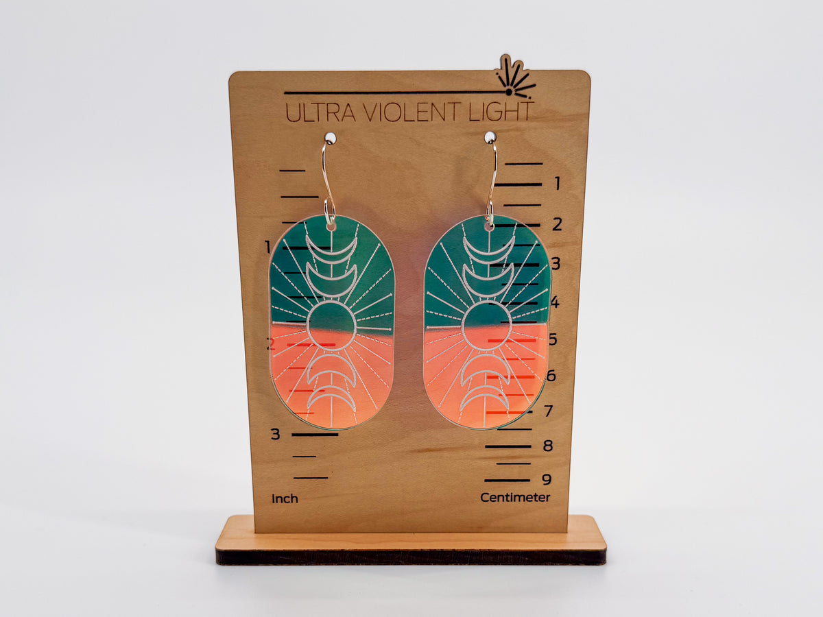 A pair of oval drop earrings displayed on a wooden stand labeled 'ULTRA VIOLENT LIGHT.' The earrings have a geometric sunburst pattern on iridescent acrylic, with silver hooks.
