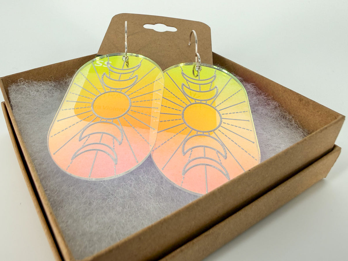 A pair of oval acrylic dangle earrings with an engraved sun and moon design and a yellow-to-pink gradient, displayed inside a brown kraft gift box with white filler on a white background.