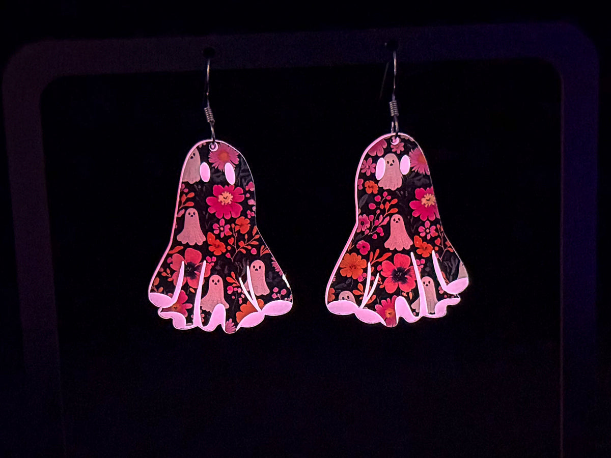 A pair of dangling earrings shaped like ghosts, decorated with a pink, red, and white floral and ghost pattern on a black background, shown glowing in the dark.