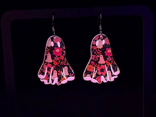 A pair of dangling earrings shaped like ghosts, decorated with a pink, red, and white floral and ghost pattern on a black background, shown glowing in the dark.
