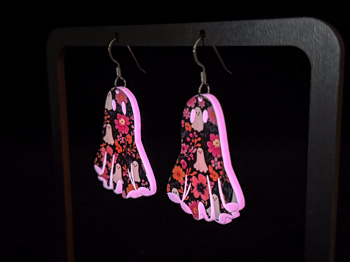 A side view of a pair of dangling earrings shaped like ghosts, decorated with a pink, red, and white floral and ghost pattern on a black background, shown glowing in the dark.
