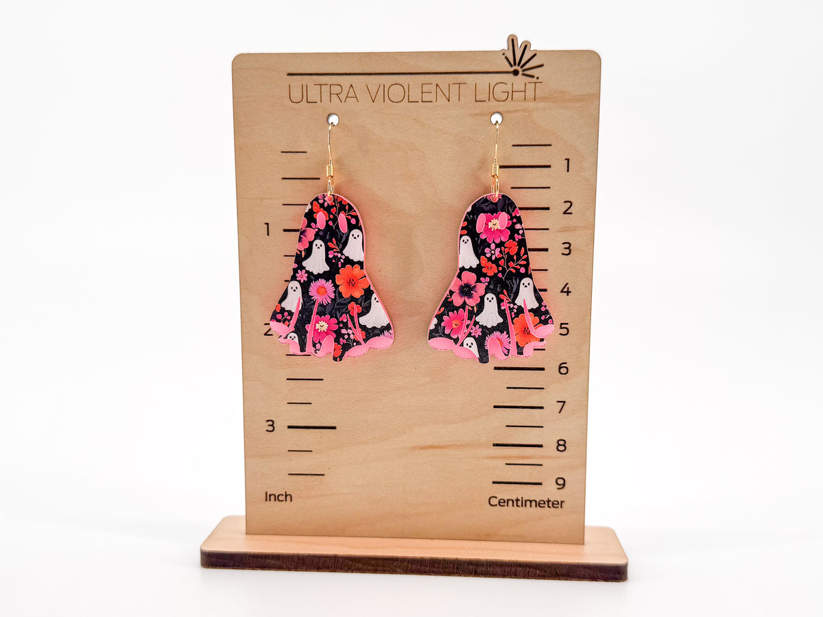 A pair of dangling earrings with a black, pink, and orange floral pattern, displayed on a wooden height-measuring stand labeled 'ULTRA VIOLENT LIGHT'.
