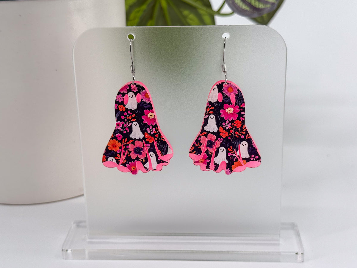 A pair of ghost-shaped dangle earrings with a pink, black, and white floral ghost pattern, displayed on a clear acrylic stand.