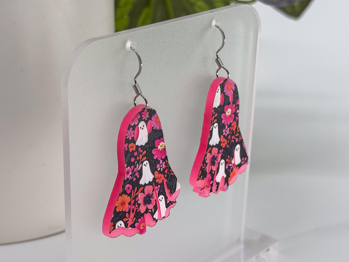 A pair of dangling earrings displayed on a clear acrylic stand. The earrings have a bright pink core and a black background covered in a floral pattern with white ghosts and small flowers.