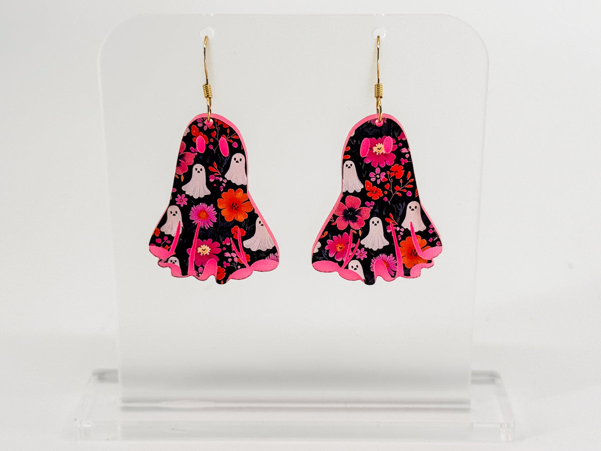 A pair of ghost-shaped drop earrings with a black background, pink and red floral pattern, white ghosts, and gold fishhook wires, displayed on a clear acrylic stand against a white background.