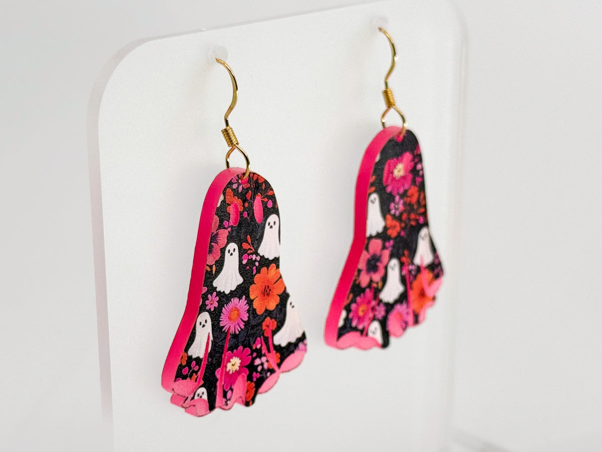A pair of dangling earrings shaped like feet, featuring a black background with a pink floral pattern, white ghost illustrations, and gold fishhook wires, displayed on a clear stand against a white background.