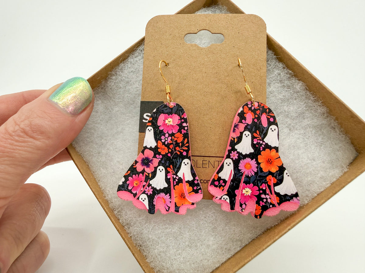 A pair of dangling earrings displayed on a brown card inside a gift box, with a hand holding the box corner. The earrings have a black background with pink, orange, and white floral and ghost patterns, attached to gold hooks.