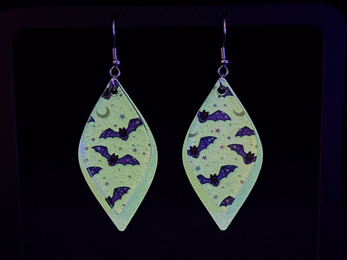 A pair of dangling teardrop-shaped earrings with a glow in the dark background, decorated with purple bats, crescent moons, and stars, shown against a black background.