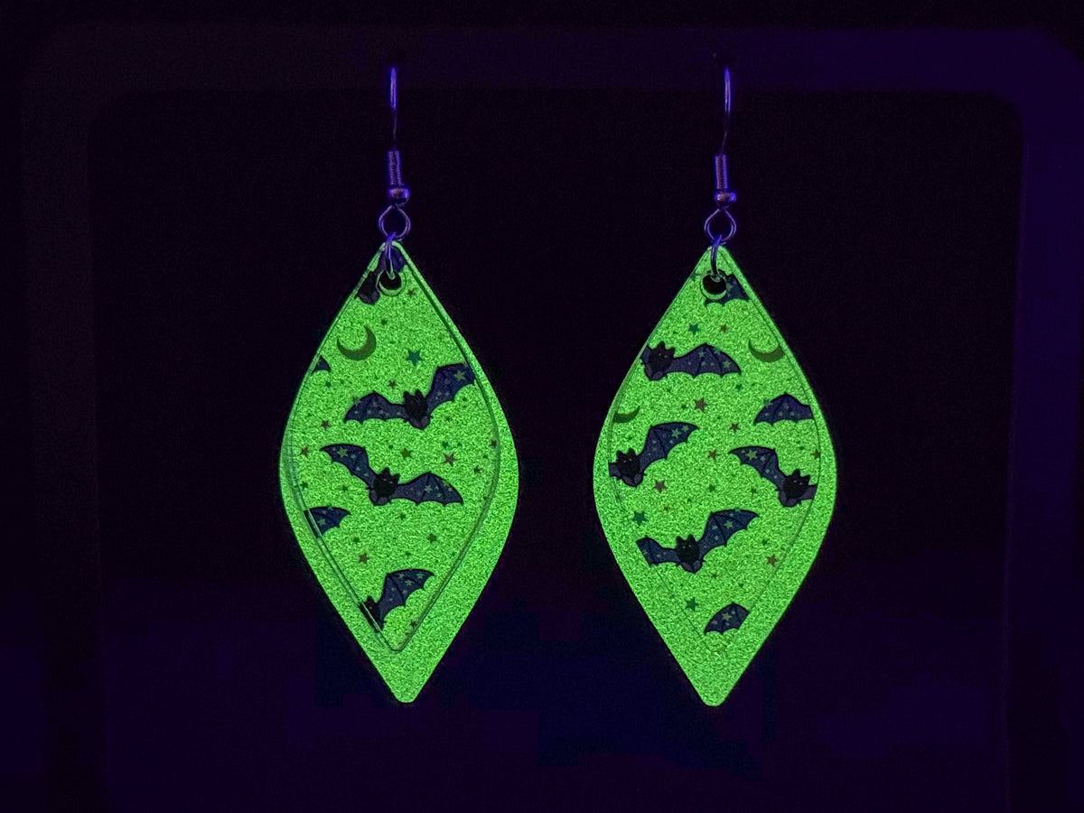 A pair of dangling teardrop-shaped earrings with a glow in the dark background and black bat and crescent moon motifs, shown against a very dark background. The glow-in-the-dark background is indeed glowing.