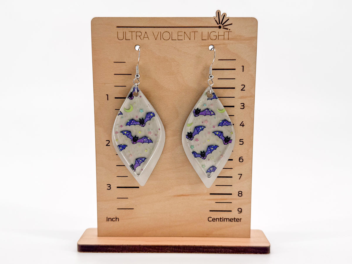 A pair of smooth rhombus-shaped earrings with purple bat designs on a cream background, displayed on a wooden height marker stand labeled 'ULTRA VIOLENT LIGHT'.