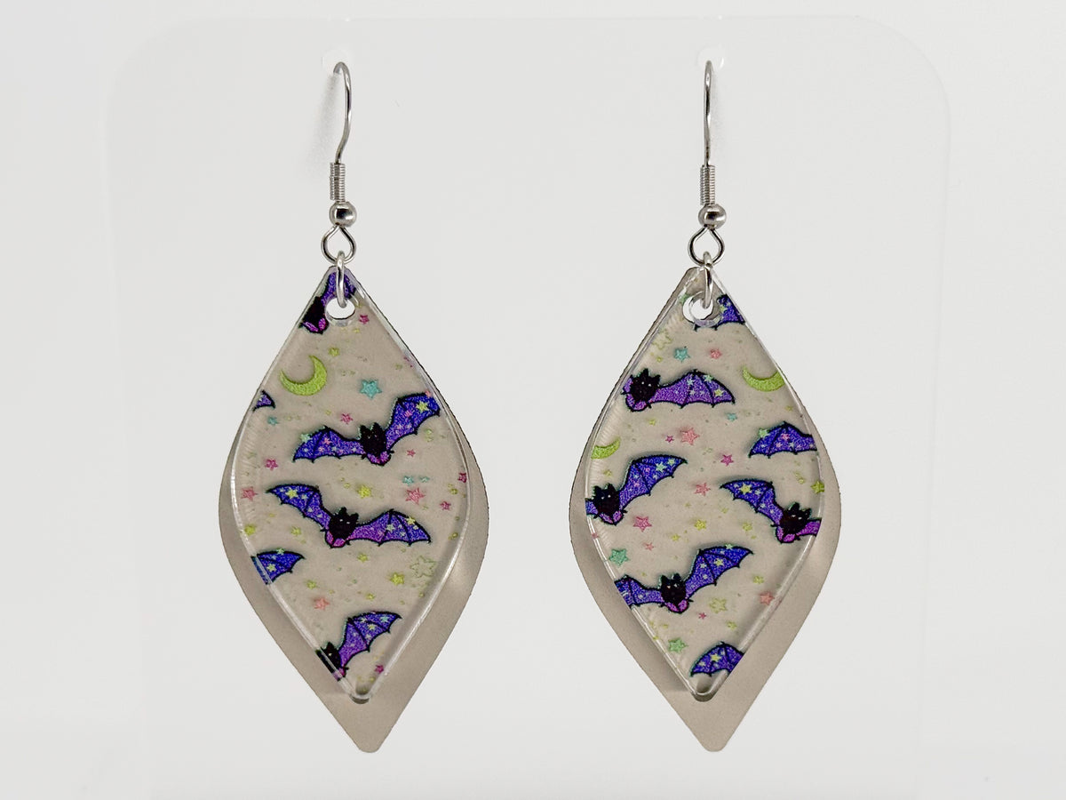 A pair of smooth rhombus-shaped dangle earrings with a silver-tone metal frame and hooks, featuring a printed pattern of purple bats, black moths, and a crescent moon on a light beige background.