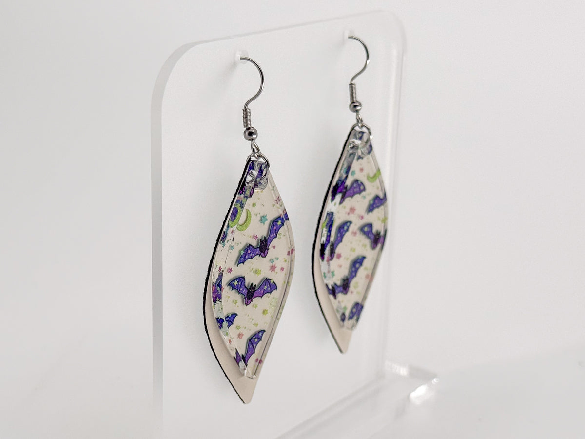 A pair of long, smooth rhombus-shaped dangle earrings with a white background and a pattern of purple, blue, and green bats and moons, displayed on a clear acrylic stand against a white background.