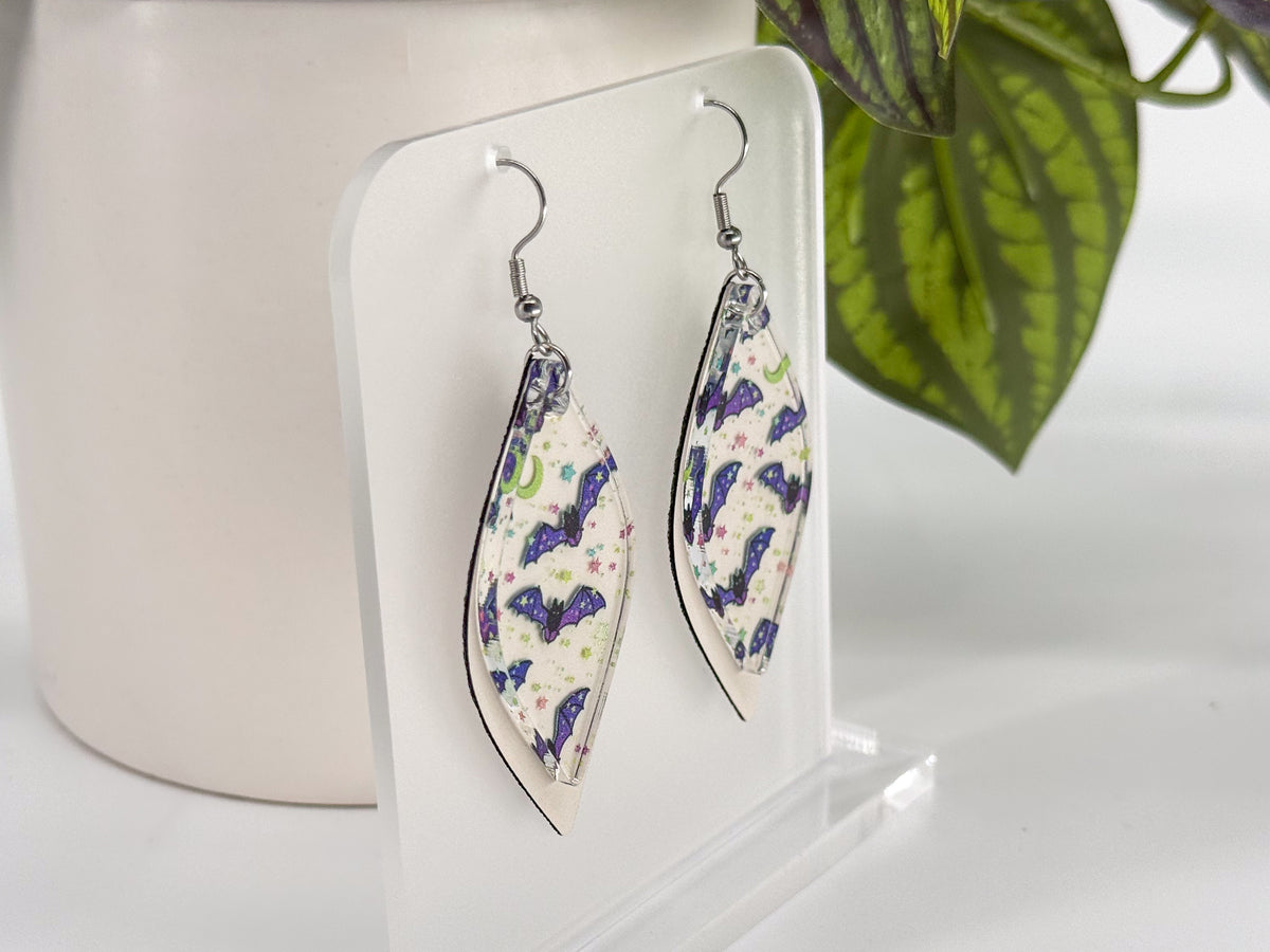 A pair of smooth rhombus-shaped dangling earrings with a colorful bat pattern on a white background, displayed on a clear acrylic stand next to a white ceramic pot and a green plant.