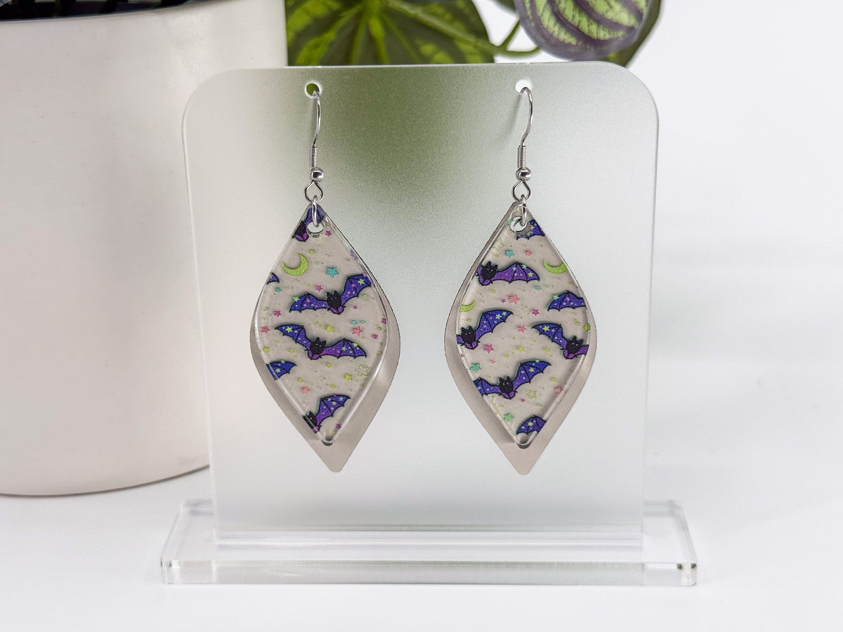 A pair of silver-tone dangle earrings with teardrop-shaped clear acrylic pendants featuring a purple bat and pink moon pattern on a white leatherette background, displayed on a clear acrylic stand.
