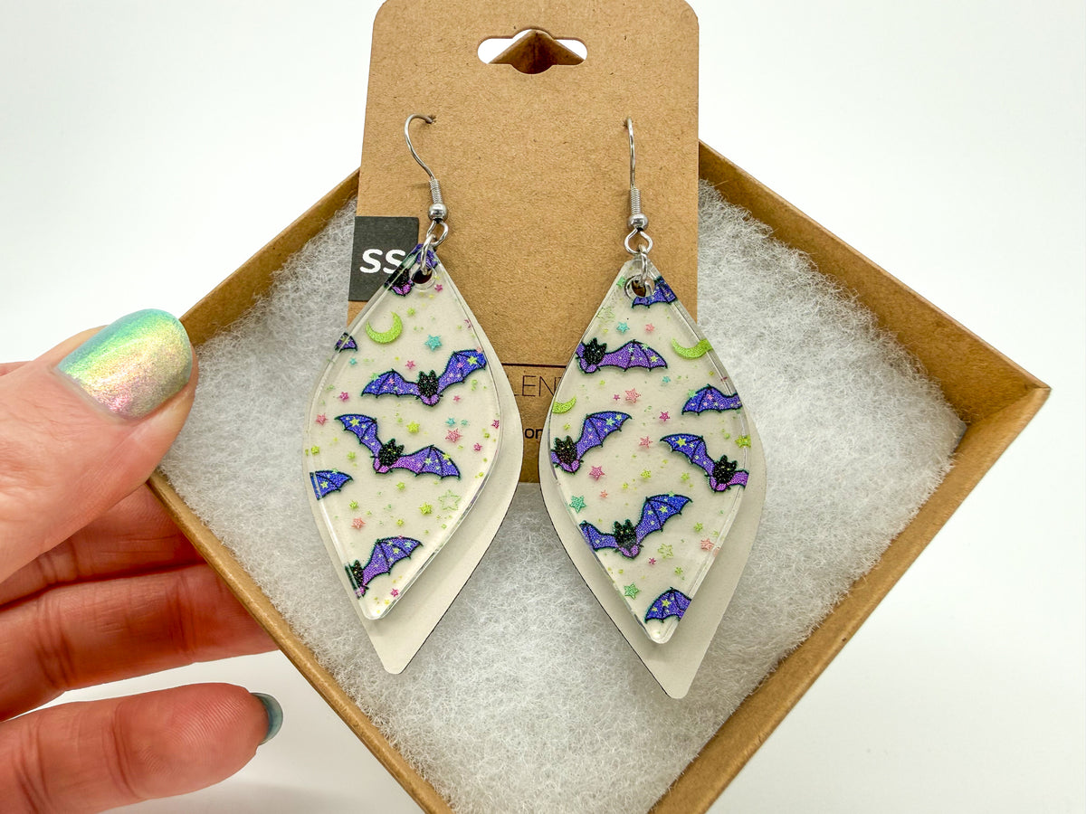 A pair of silver-tone dangle earrings with cream-colored smooth rhombus-shaped charms printed with purple bats, black wings, and small pink and green details, held by a hand in front of a brown gift box.