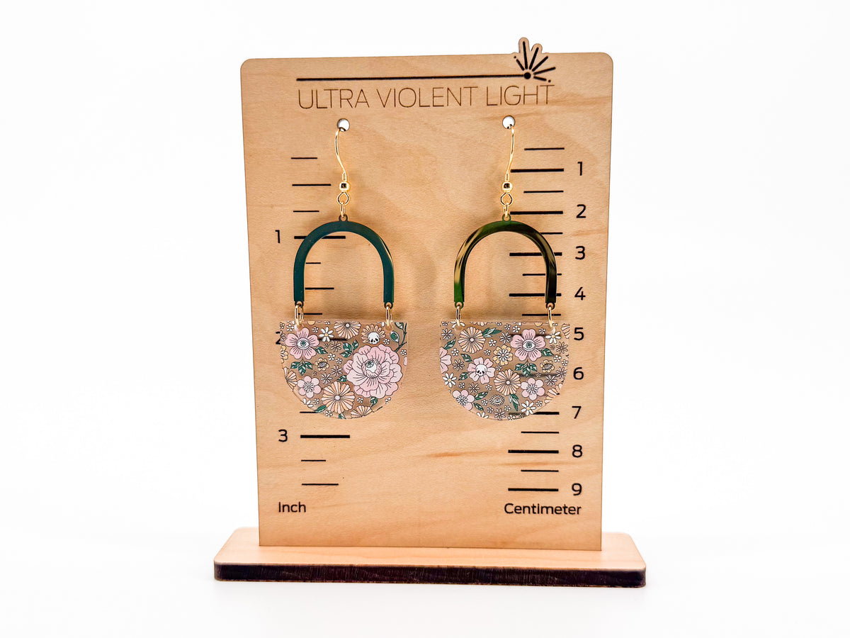 A pair of statement earrings displayed on a wooden height-measuring stand against a white background. The earrings feature gold arches with floral designs in pink, white, and green.