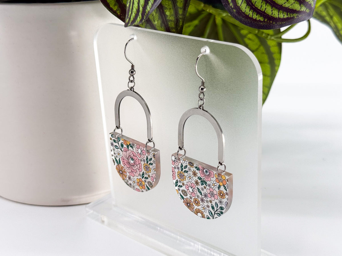 A pair of dangling earrings displayed on a clear acrylic stand. Each earring has a silver-tone arch top and a curved lower section covered in a dense multicolor floral pattern with pink, orange, yellow, green, and blue tones, featuring hidden skulls and eyes.