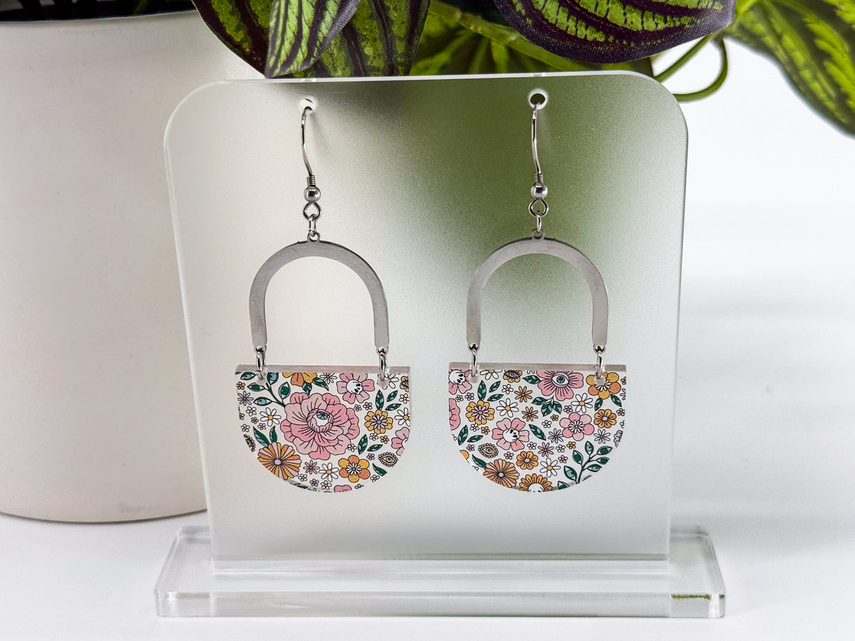 A pair of dangling earrings displayed on a clear acrylic stand. Each earring has a silver-tone arch top and a curved lower section covered in a dense multicolor floral pattern with pink, orange, yellow, green, and blue tones, featuring hidden skulls and eyes.