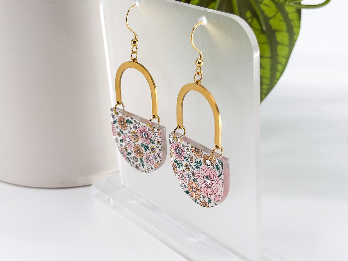 A pair of dangling earrings displayed on a clear acrylic stand. Each earring has a gold-tone arch top and a curved lower section covered in a dense multicolor floral pattern with pink, orange, yellow, green, and blue tones, featuring hidden skulls and eyes.