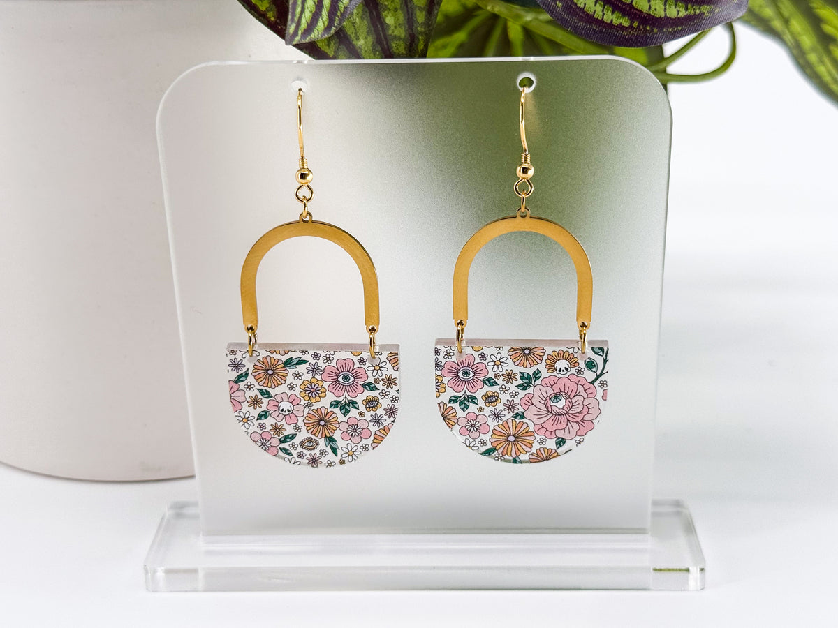 A pair of dangling earrings displayed on a clear acrylic stand. Each earring has a gold-tone arch and hook with a small floral-patterned half-circle below (and hidden skulls and eyeballs).