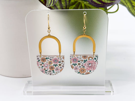 A pair of dangling earrings displayed on a clear acrylic stand. Each earring has a gold-tone arch and hook with a small floral-patterned half-circle below (and hidden skulls and eyeballs).