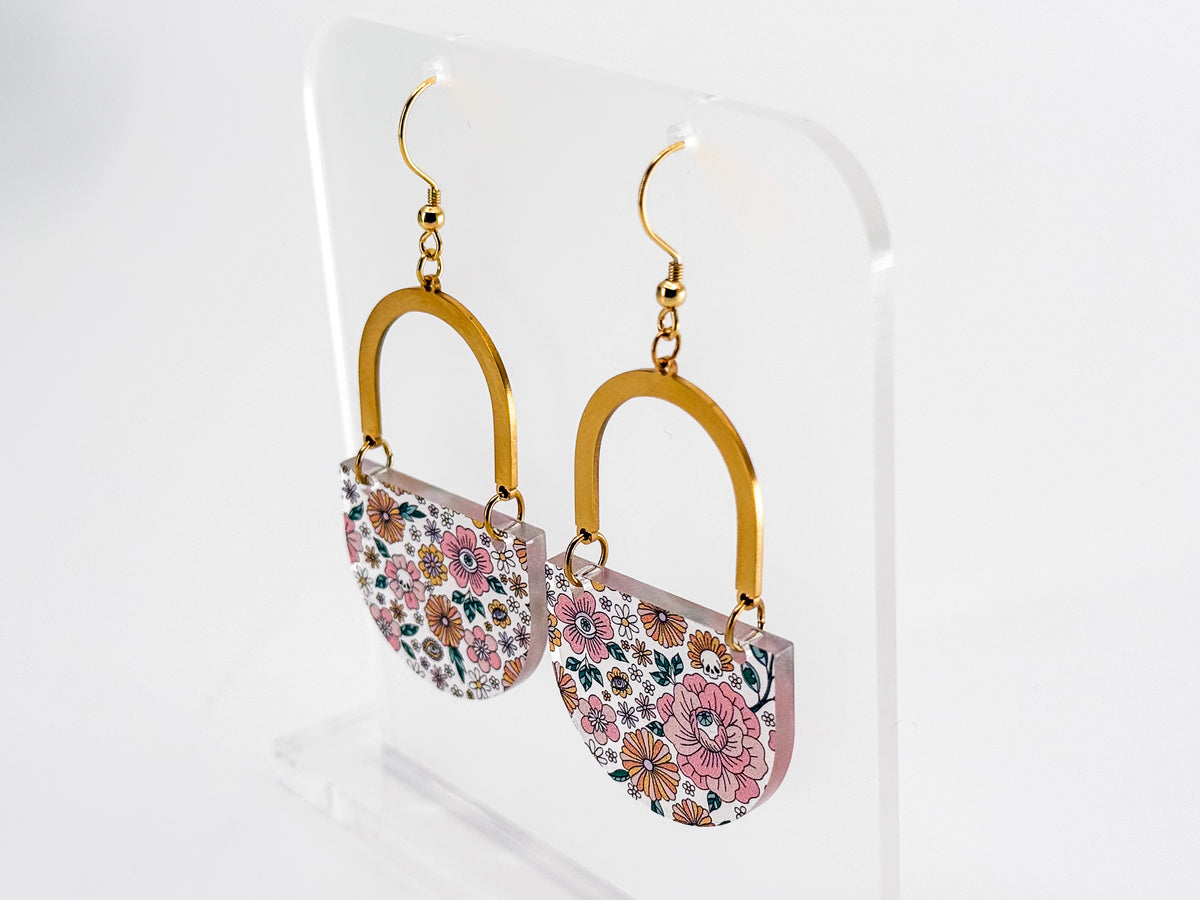 A pair of dangling earrings displayed on a clear acrylic stand. Each earring has a gold-tone arch top and a curved lower section covered in a dense multicolor floral pattern with pink, orange, yellow, green, and blue tones, featuring hidden skulls and eyes.