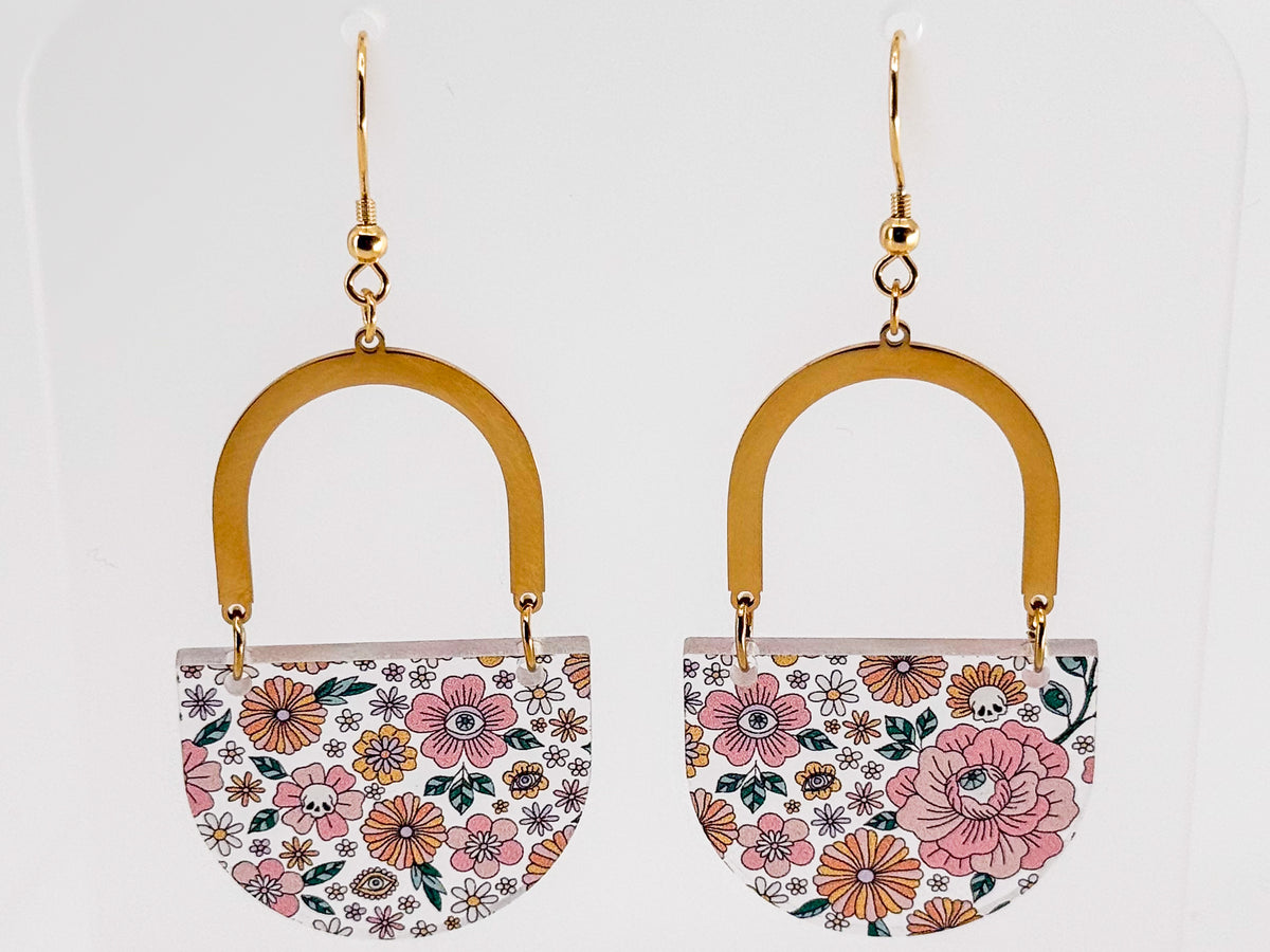 A pair of dangling earrings with gold-tone hooks and arches, featuring semi-circular floral panels with pink, orange, and white flowers (and hidden skulls and eyeballs) on a white background.