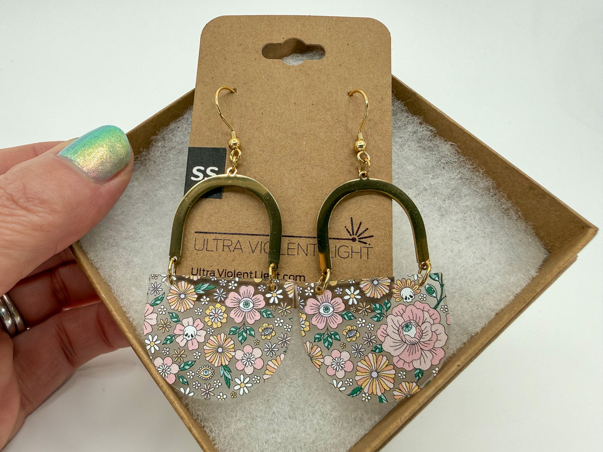 A pair of floral hoop drop earrings with gold-tone hoops and floral charms, displayed on a brown card inside a box, with a hand holding the left side.