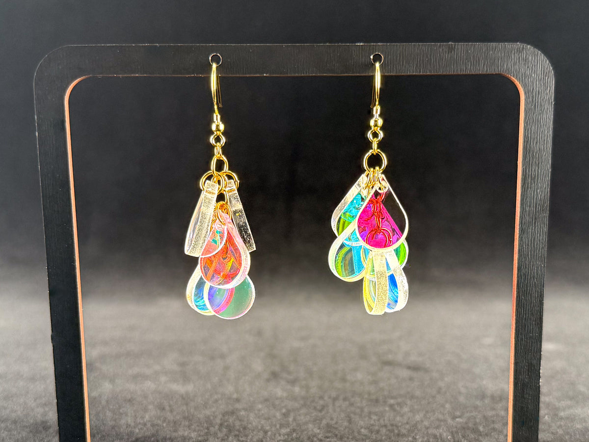 A pair of dangling earrings displayed on a black stand against a dark background. Each earring has a gold-tone hook and a cluster of iridescent, teardrop-shaped charms that reflect pink, blue, green, and yellow tones.