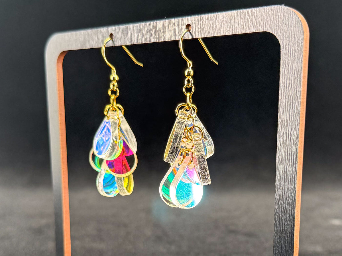 A pair of gold-tone dangle earrings with translucent iridescent teardrop charms, displayed on a black stand with a wooden frame against a dark background.