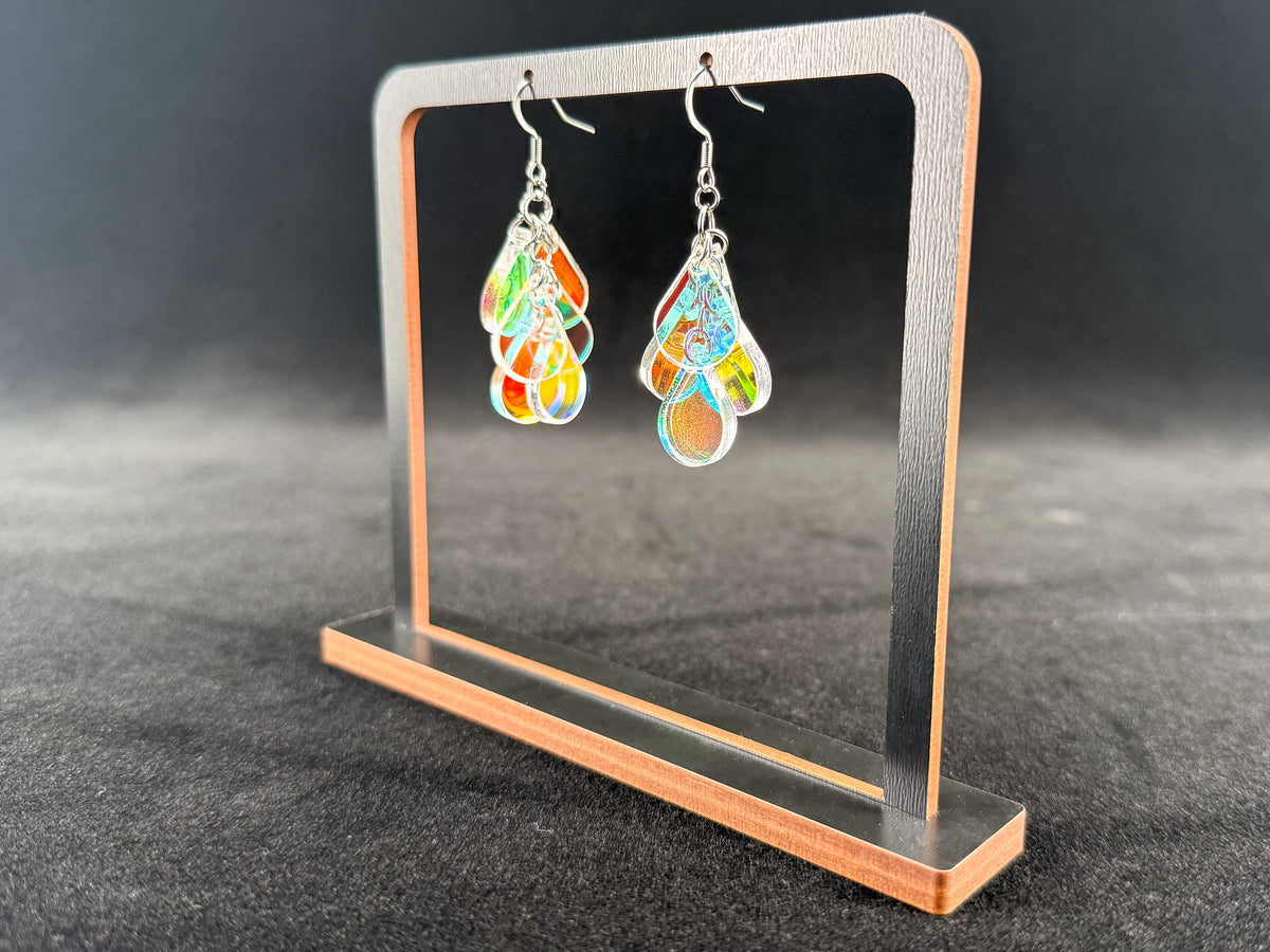 A pair of colorful dangling earrings displayed on a small rectangular stand against a dark gray background. The earrings have a silver-tone metal frame with layered teardrop-shaped acrylic charms in orange, yellow, green, and blue.