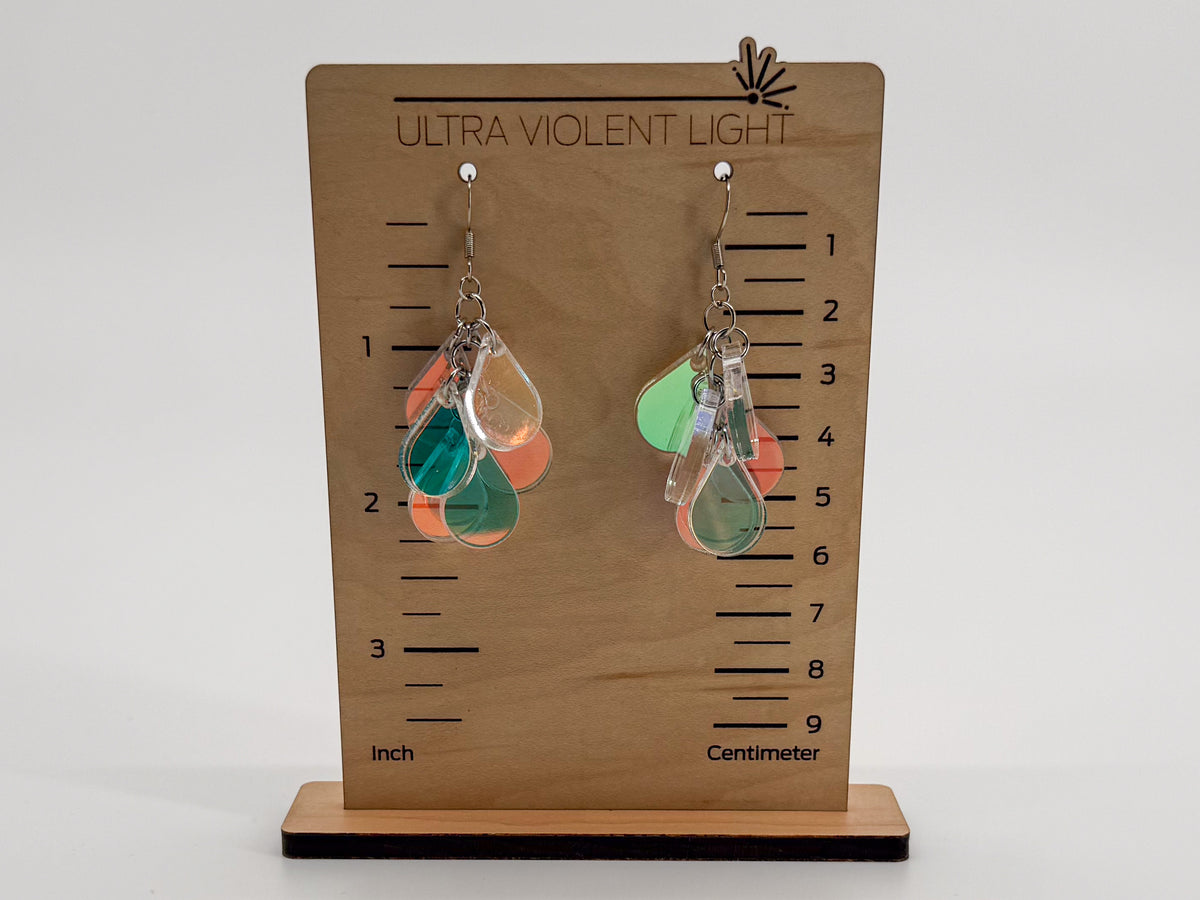 A pair of dangling teardrop-shaped earrings displayed on a wooden height-measuring stand labeled 'ULTRA VIOLENT LIGHT.' The earrings appear translucent with overlapping green, orange, and clear sections, and the stand includes inch and centimeter markings.