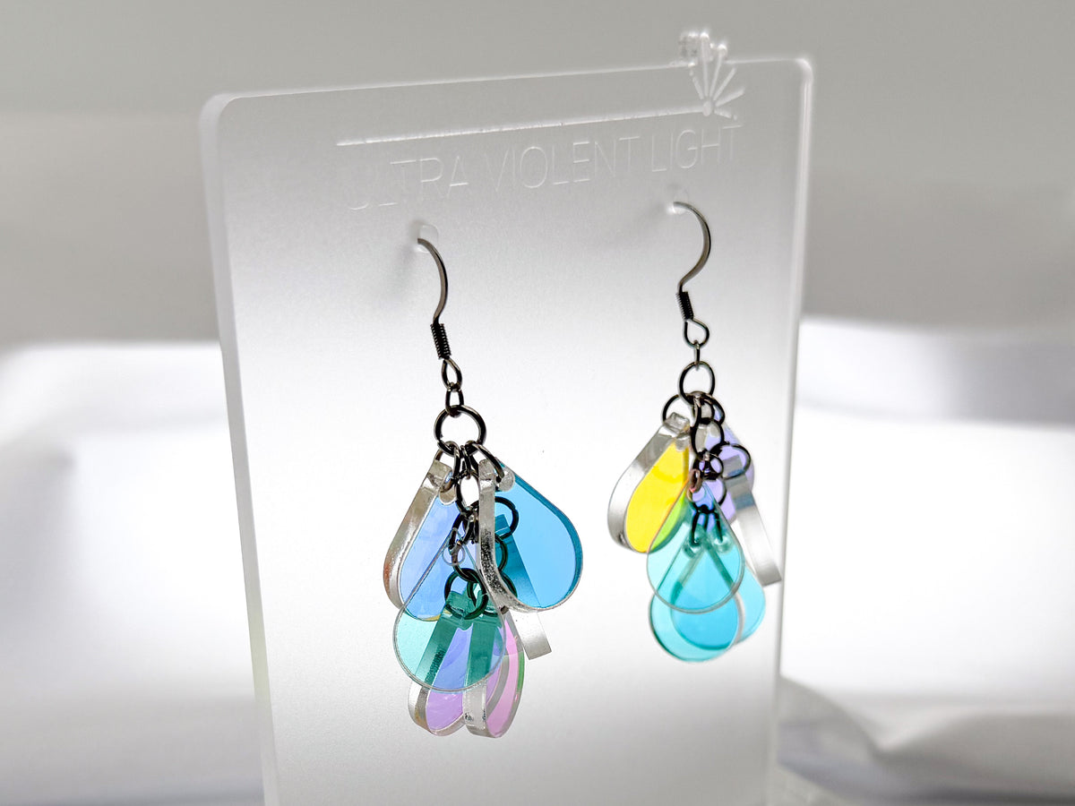 A pair of dangling earrings displayed on a clear acrylic stand. The earrings feature multiple iridescent teardrop-shaped acrylic charms in blue, green, purple, and yellow, suspended from silver hooks.