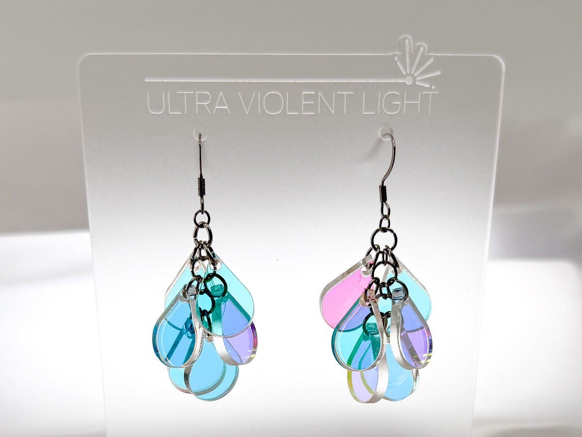 A pair of dangling earrings displayed on a clear acrylic stand labeled 'ULTRA VIOLENT LIGHT.' The earrings have silver-toned hooks and clustered teardrop-shaped iridescent pieces in light blue, aqua, lavender, and pink, photographed against a soft white background.