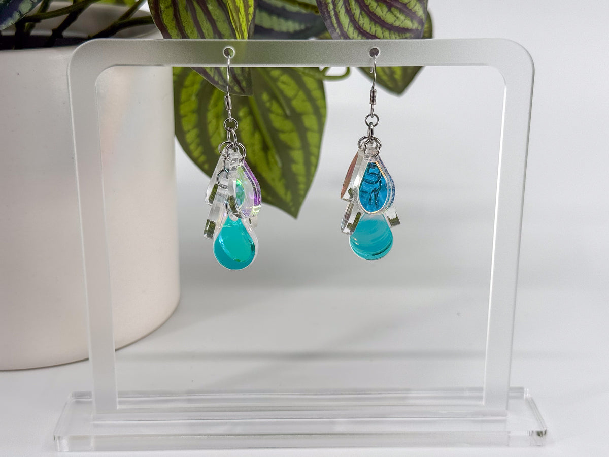 A pair of dangling earrings displayed on a clear acrylic stand. Each earring has a silver-toned hook iridescent acrylic teardrop shapes below it, set against a white background with green leaves in the upper background.