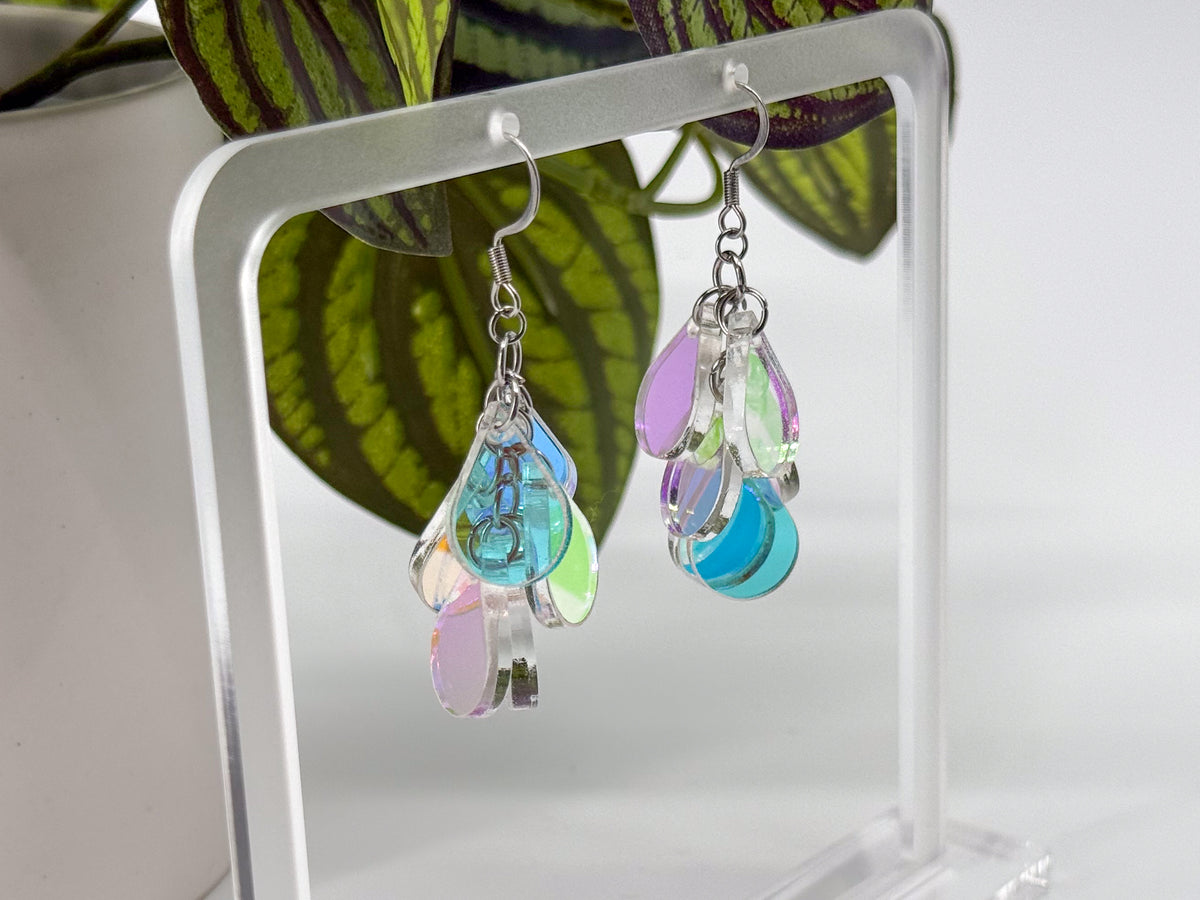 A pair of dangling earrings with silver-toned hooks and iridescent teardrop-shaped charms in pastel blue, pink, green, and lavender, displayed on a clear acrylic stand in front of green leaves.