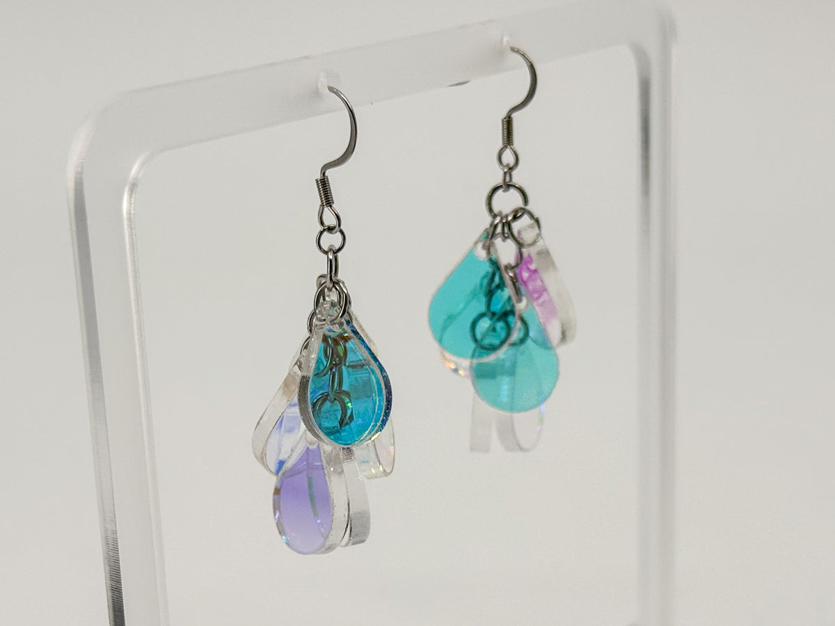 A pair of handmade teardrop dangle earrings displayed on a clear acrylic stand. The earrings feature silver-toned metal hooks and multiple dangling clear acrylic charms with blue, purple, and teal colors.