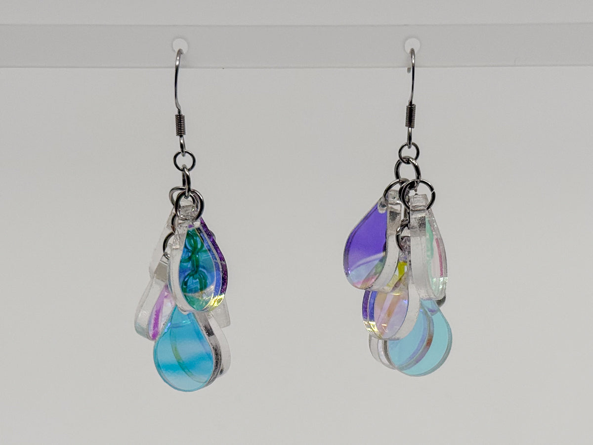 A pair of dangling earrings with multiple iridescent teardrop-shaped charms in blue, purple, green, and pink, suspended from silver hooks against a plain light gray background.