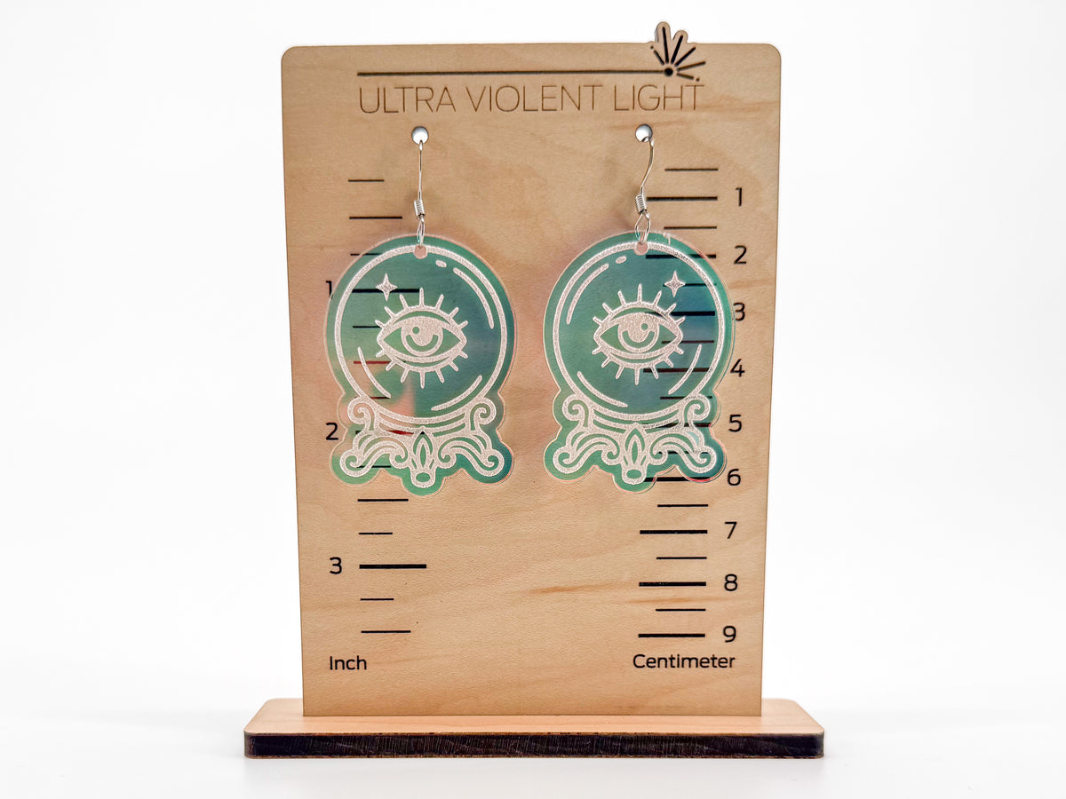 A pair of dangling earrings displayed on a wooden measurement stand. The earrings are circular with an iridescent engraved eye design and silver hooks.