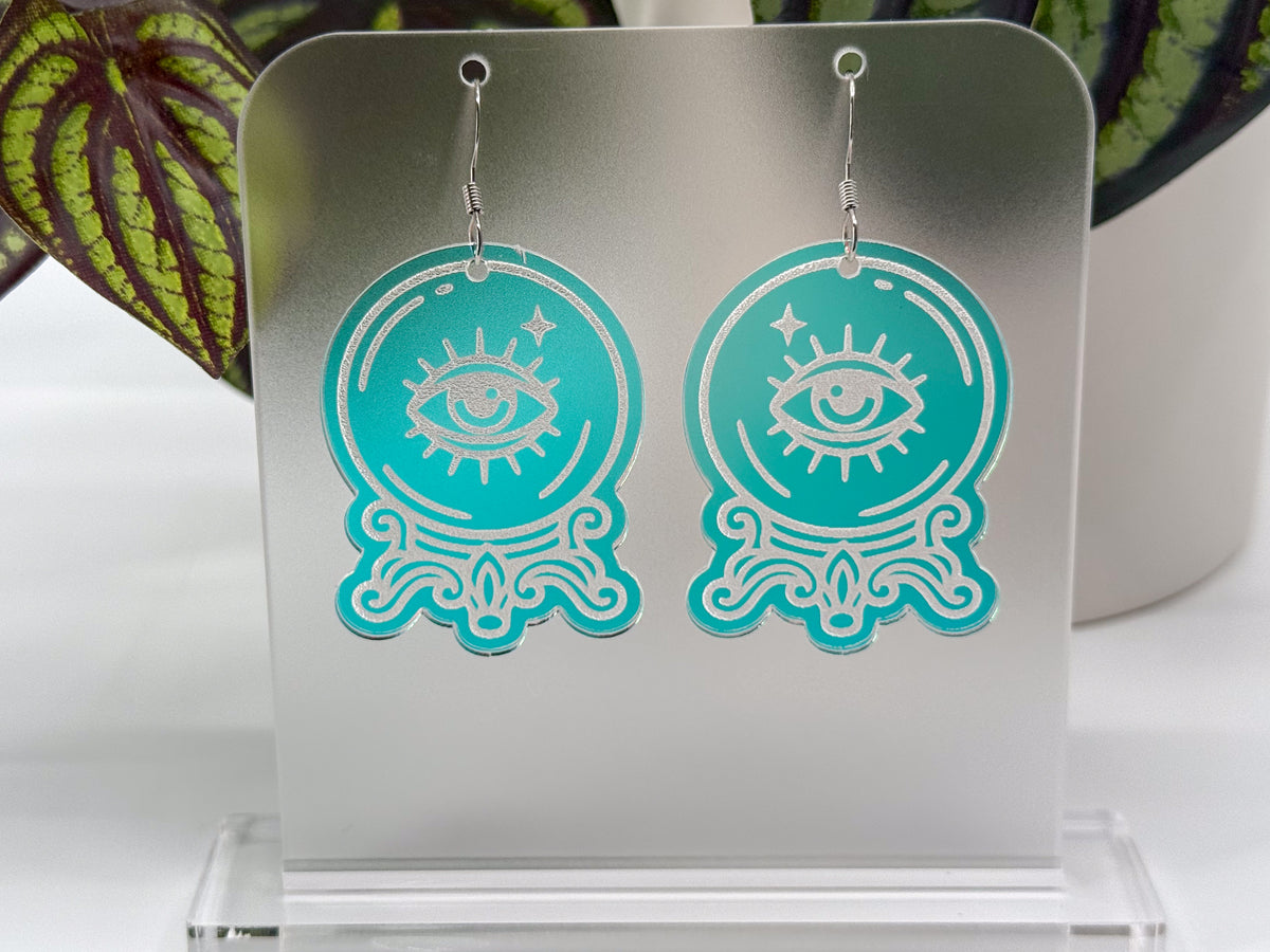 A pair of iridescent laser-cut acrylic dangle earrings with an eye-in-a-crystal ball design, displayed on a clear acrylic stand.