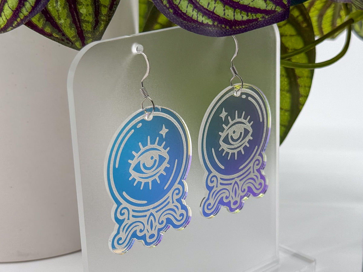 A pair of oval acrylic dangle earrings with a white all-seeing eye and star design, featuring a blue-to-purple gradient and holographic shimmer, displayed on a clear acrylic stand.
