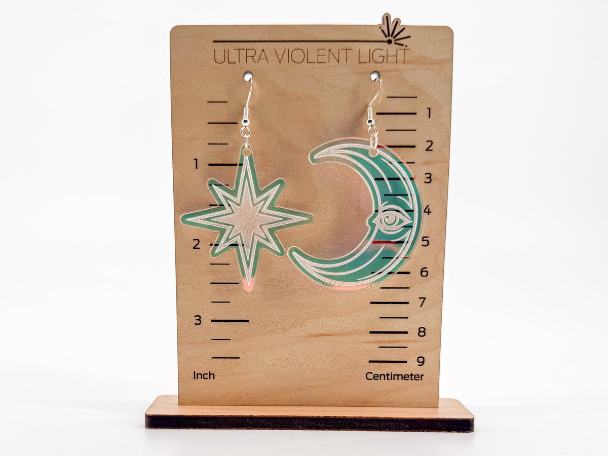 A pair of dangling earrings displayed on a wooden measurement stand labeled 'ULTRA VIOLENT LIGHT.' The earrings are  crescent moon and star shapes in iridescent acrylic.
