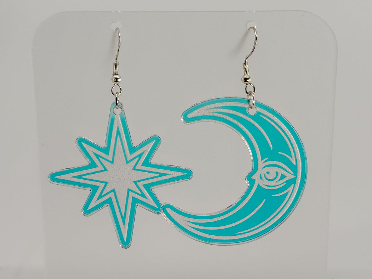 A pair of dangling earrings displayed on a clear acrylic stand. One earring is a turquoise and white star, and the other is a crescent moon with a white eye design.