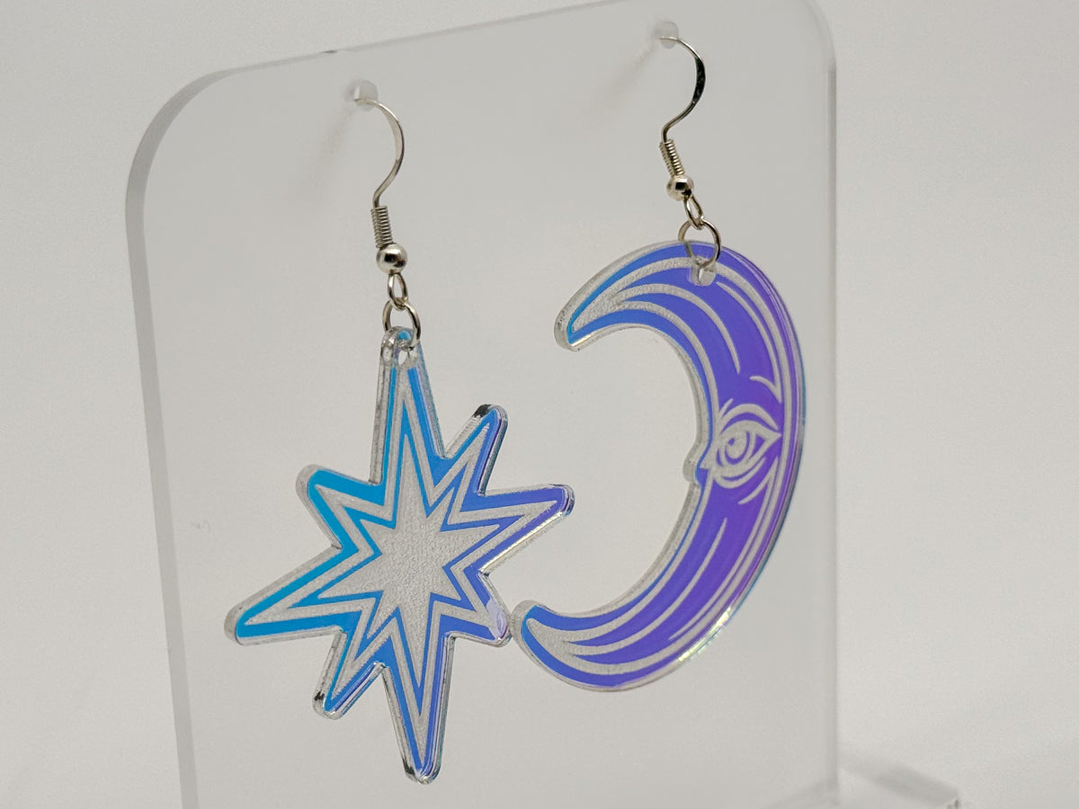 A pair of dangling earrings displayed on a clear acrylic stand. One earring is a blue outlined star, and the other is a purple outlined crescent moon with a stylized eye design.