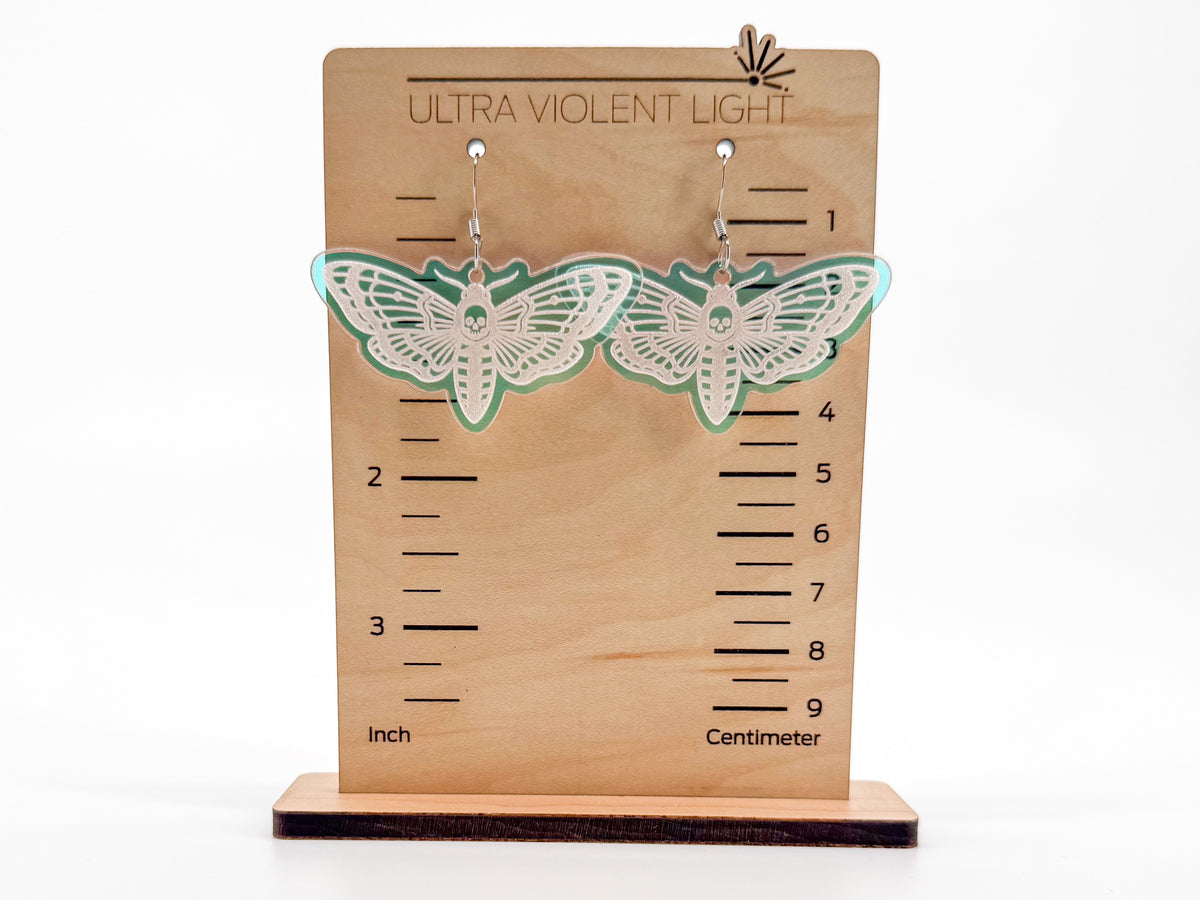 A pair of moth-shaped earrings of translucent iridescent acrylic, with silver-toned hooks, displayed on a wooden height-measuring stand labeled 'ULTRA VIOLENT LIGHT'.
